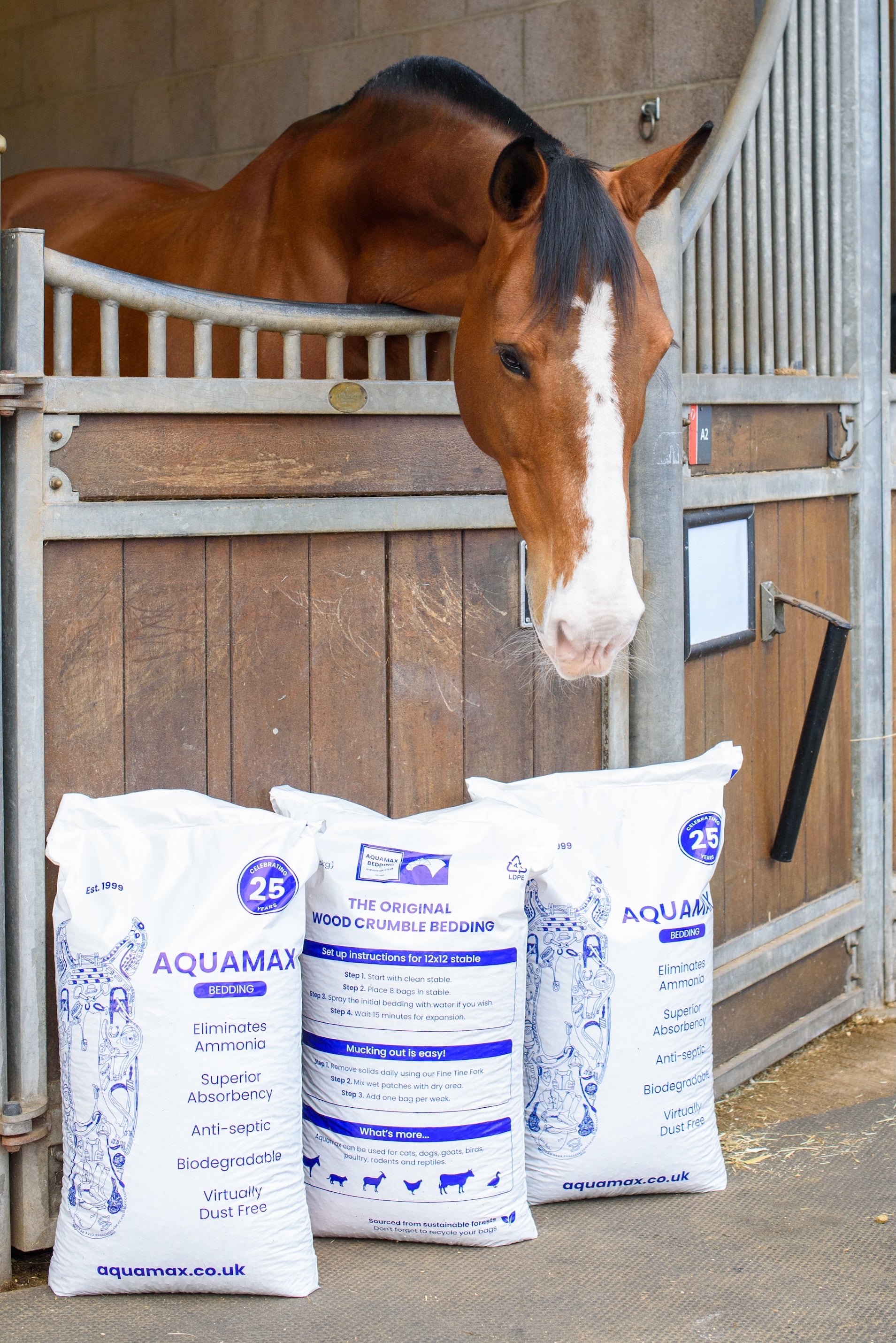 Three bags of Aquamax horse bedding pellets are placed on the ground with a horse looking over a stable door in the background.